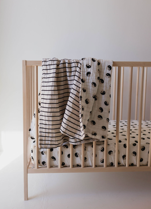 A light-colored wooden crib features bedding and two baby blankets draped over the side. The fitted sheet and one blanket have a pattern of black, celestial crescent moons, while the other blanket is the "Muslin Quilt | Black & White Yin Yang / Stripe" by forever french baby, made of 100% muslin cotton with black horizontal stripes on a white background.