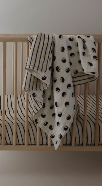 A light wooden crib with a striped mattress and two blankets hanging over the side, including a Black & White Yin Yang quilt and a reversible 100% muslin cotton quilt from forever french baby, featuring black stripes on one side.