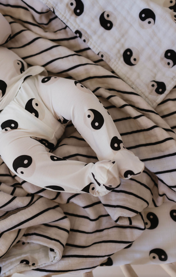 A baby, dressed in white clothing adorned with black yin-yang symbols, lies on a reversible Muslin Quilt | Black & White Yin Yang / Stripe from forever french baby. This quilt, made from 100% muslin cotton, features the same yin-yang pattern and black stripes beneath the baby.