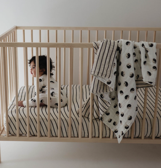 A young child sits in a wooden crib, dressed in a white outfit with small black designs. The crib features a striped mattress and two blankets draped over the side: one with black stripes and the other being the Muslin Quilt | Black & White Yin Yang / Stripe by forever french baby, made from 100% muslin cotton. The background is plain and light-colored.