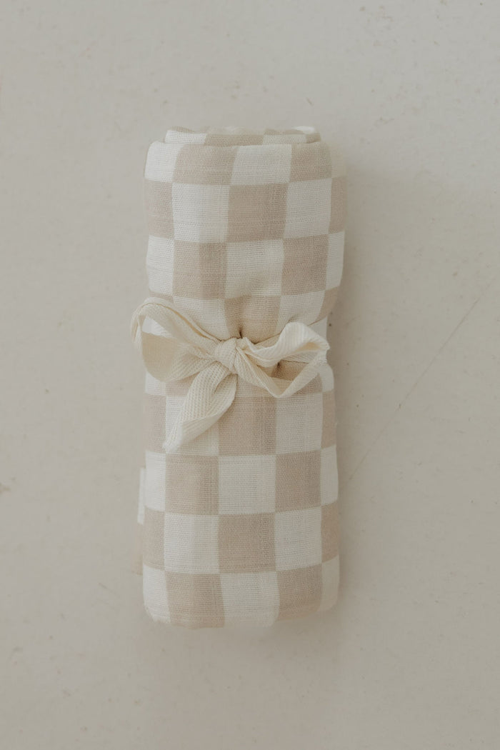 The Muslin Swaddle | Checkerboard by forever french baby, featuring a beige and white gingham pattern, is rolled up and tied with a cream ribbon, displayed on a light, textured surface.