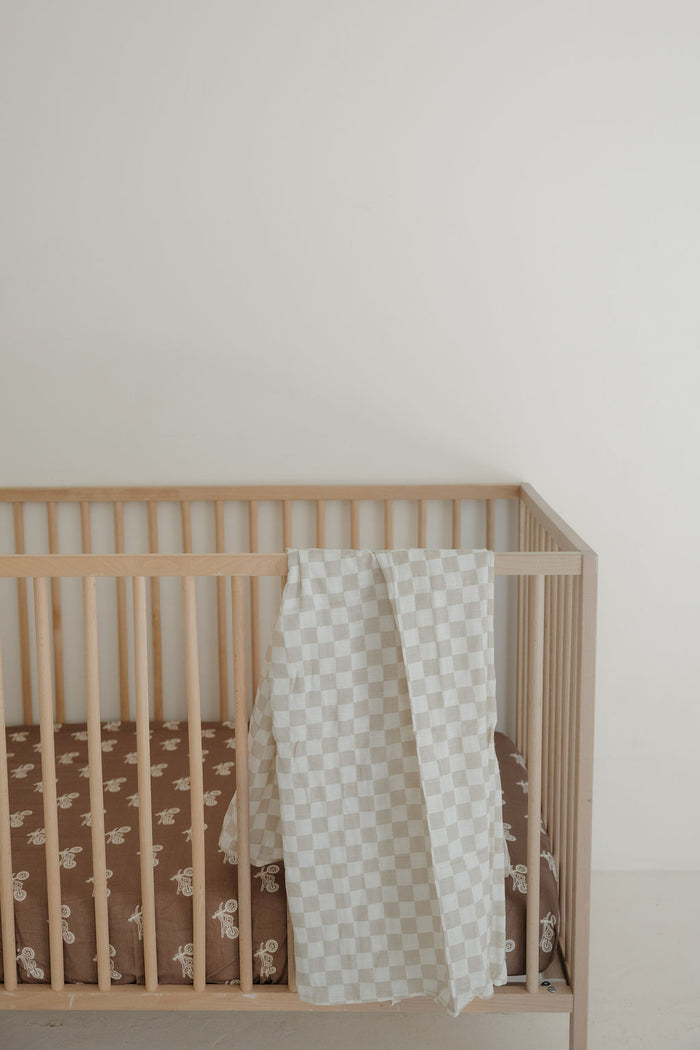 A light wooden crib with a brown mattress featuring white patterns and a forever french baby Muslin Swaddle | Checkerboard draped over the side, set against a plain white wall.