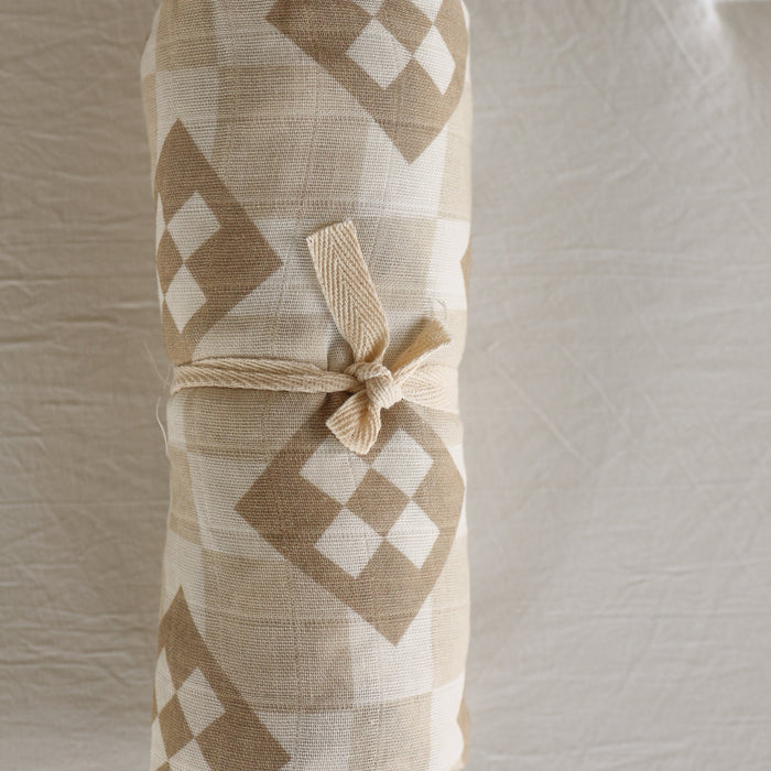 The forever french baby Muslin Swaddle | Cottage Quilt features a beige and white geometric diamond pattern, secured with a knotted beige tie, and is displayed on a textured light background.