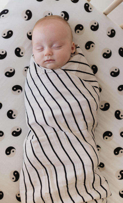 A baby wrapped tightly in a Muslin Swaddle | Grey & Black Stripe by forever french baby sleeps peacefully. The little one lies on a fitted sheet featuring a black and white yin-yang pattern, creating a serene scene that exudes calmness and tranquility.