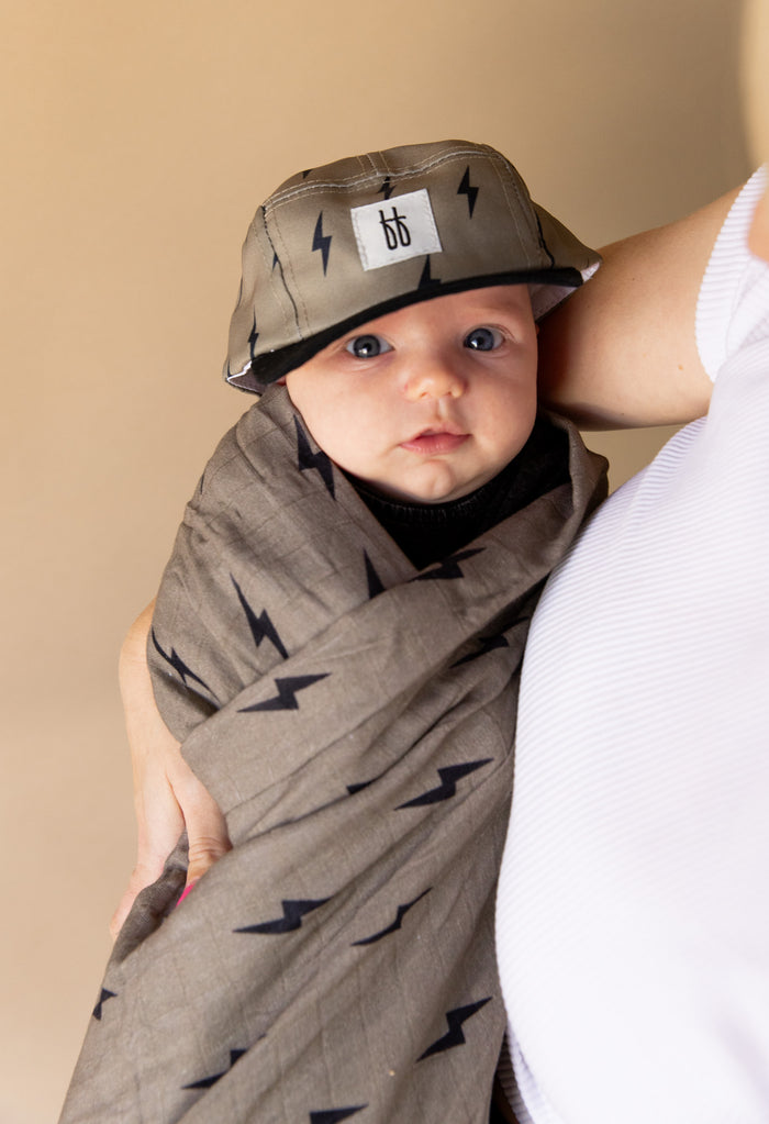 A baby, cozily wrapped in the "Muslin Swaddle | Lightning Bolt" from forever french baby, is being held by an adult. The baby also sports a gray cap adorned with black lightning bolts and a white "bb" logo. Meanwhile, the adult is dressed in a white top against a beige background.