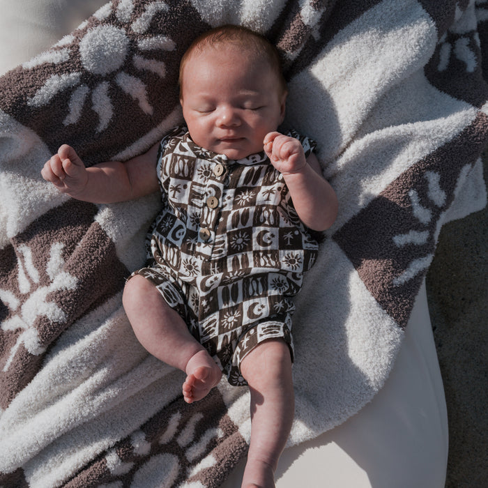 A baby in a forever french baby Muslin Tank Romper | Summer Check lies on a sun and moon-themed blanket outdoors, eyes closed and arms raised, relaxing in natural light.