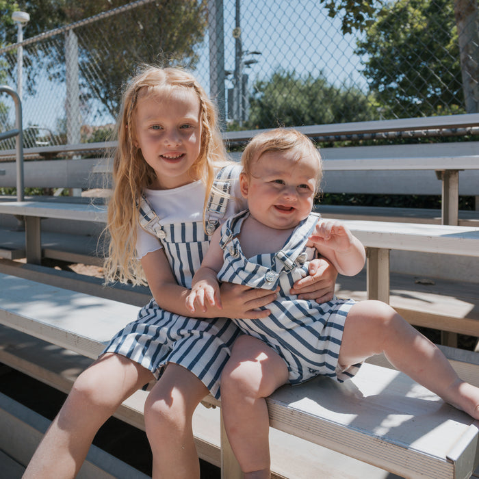 Two young children sit outdoors on bleachers. The older child smiles while holding a baby; both wear matching "Denim Stripe" overalls from forever french baby. Sunlight gently filters through, with a fence and trees seen in the background.