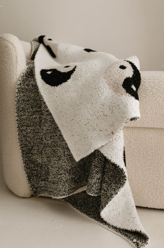 A plush blanket named "Black & White Yin Yang" by Forever French Baby, with a velvety-rich texture, drapes over the arm of a cream-colored, textured chair. The blanket showcases a stylized panda face with black ears, eyes, and nose on a white background. The scene exudes a cozy and minimalist aesthetic.