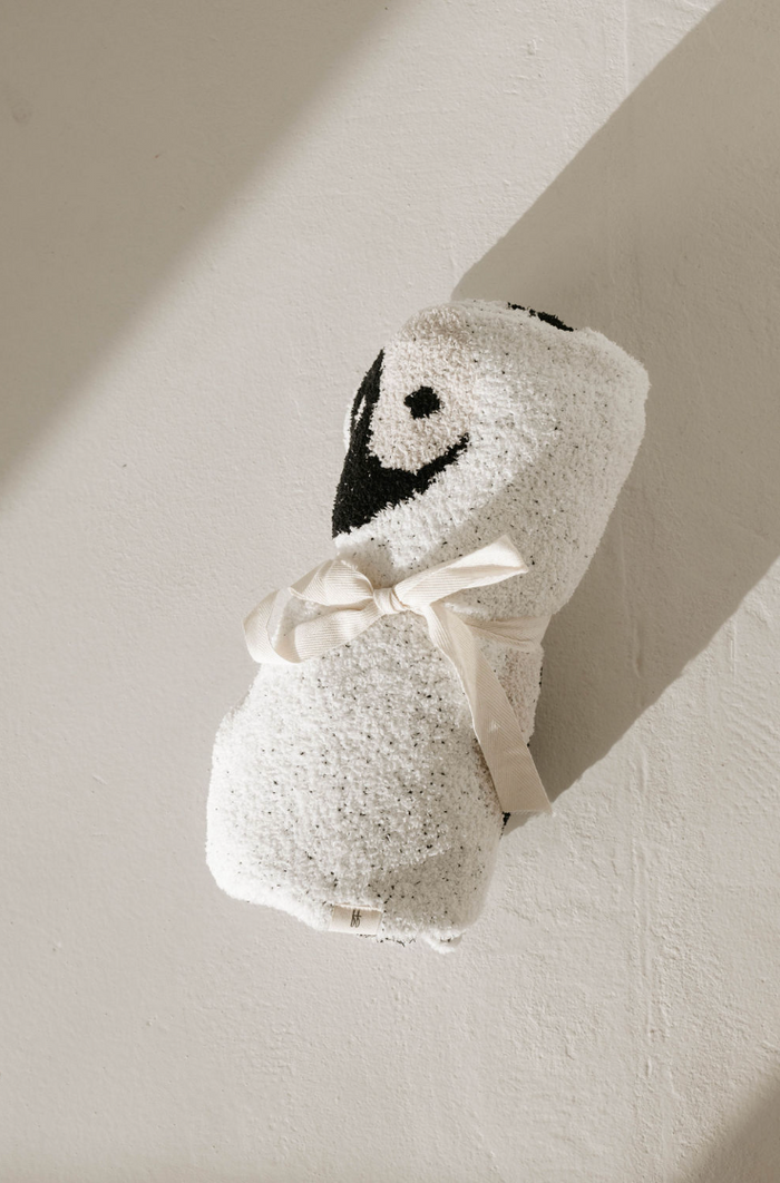 A Plush Blanket | Black & White Yin Yang from forever french baby, textured and velvety-rich, is rolled and tied with a beige ribbon on a light surface. Made from microfiber feather yarn, it features a black and white embroidered penguin face near one end, adding a playful touch to the luxurious bundle. Shadows fall on the surface, creating depth.