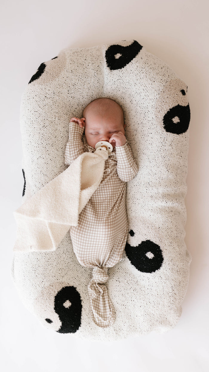 A baby sleeps on a white, pillow-like cushion decorated with black yin-yang symbols from the forever french baby Plush Blanket | Black & White Yin Yang collection. The baby is wearing a beige and white gingham onesie made from OEKO-TEX Standard 100 fabric and holds a beige pacifier while grasping a soft, light-colored cloth. The baby has a peaceful expression with one hand raised.