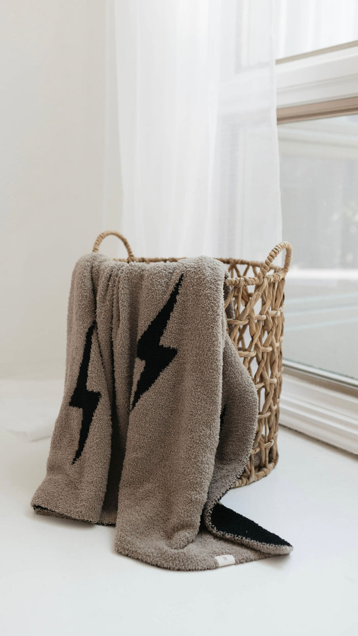 A woven basket with handles rests on a white surface by a large window. A Plush Blanket from forever french baby, featuring brown lightning bolt designs and made of Microfiber feather yarn, hangs partially out of the basket. Sheer white curtains are drawn to the side, allowing natural light to fill the space.