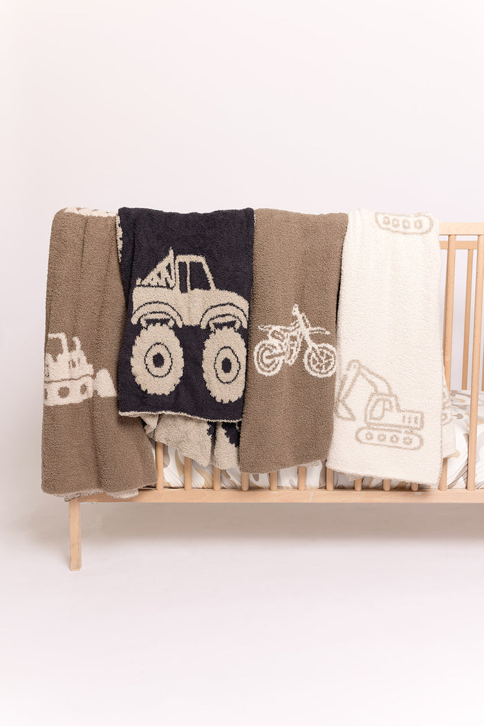 Three forever french baby Plush Blankets | Bulldozer in brown, cream, and black tones with construction vehicle and motorcycle designs are draped over a light wooden crib against a plain white background.
