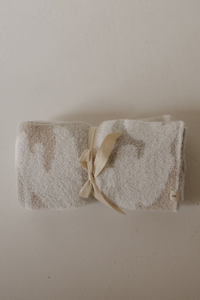A cozy, OEKO-TEX certified Plush Blanket | Camo 2.0 by forever french baby, featuring a beige and white pattern, is neatly rolled and tied with a cream ribbon, resting on a light textured surface.