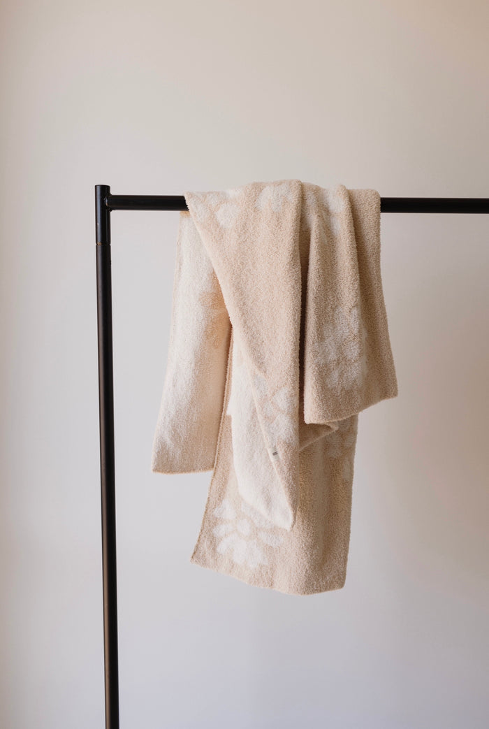 A Lazy Daisy Plush Blanket from forever french baby, featuring a subtle floral pattern in beige, made from velvety-rich microfiber feather yarn, is draped over a minimalist black metal towel rack. The background is a plain, light-colored wall, giving a clean and simple aesthetic to the image. This luxurious blanket meets the OEKO Standard 100.