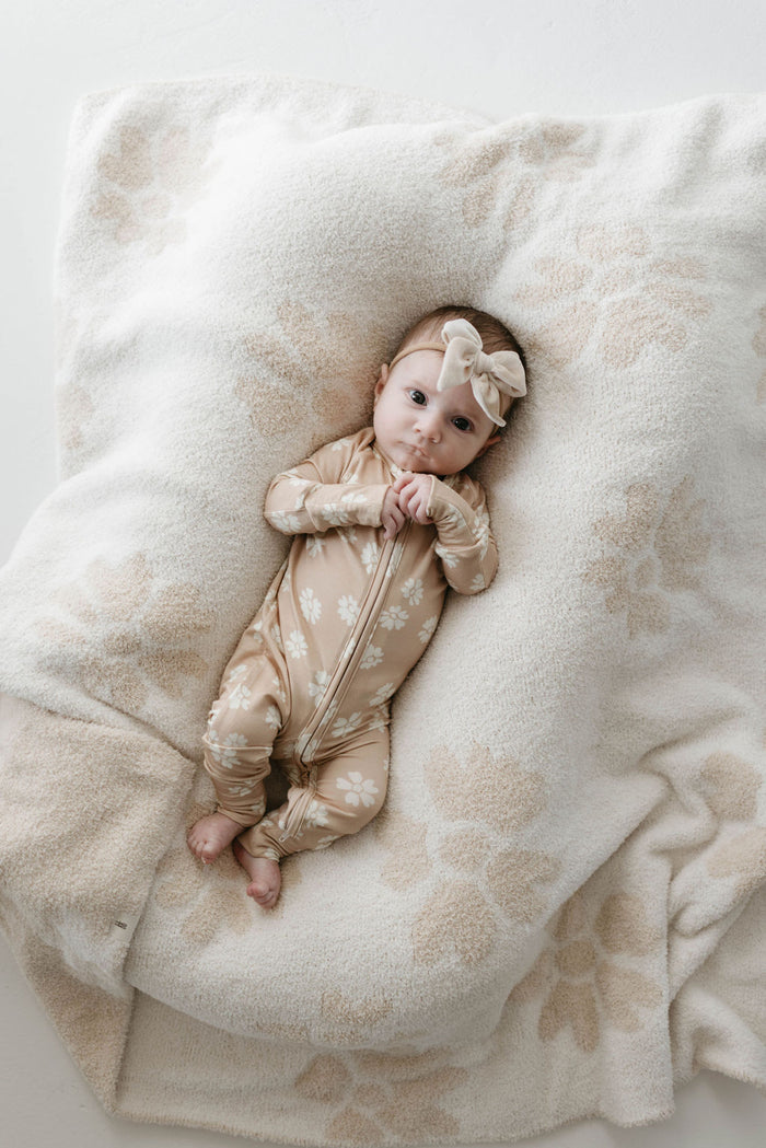 A baby is lying on the Lazy Daisy Plush Blanket from forever french baby, which features a fluffy white and beige floral design made from OEKO Standard 100 materials. The baby is wearing a beige onesie adorned with white floral patterns and has a matching beige bow headband. The baby gazes upwards while touching the velvety-rich texture of the blanket.