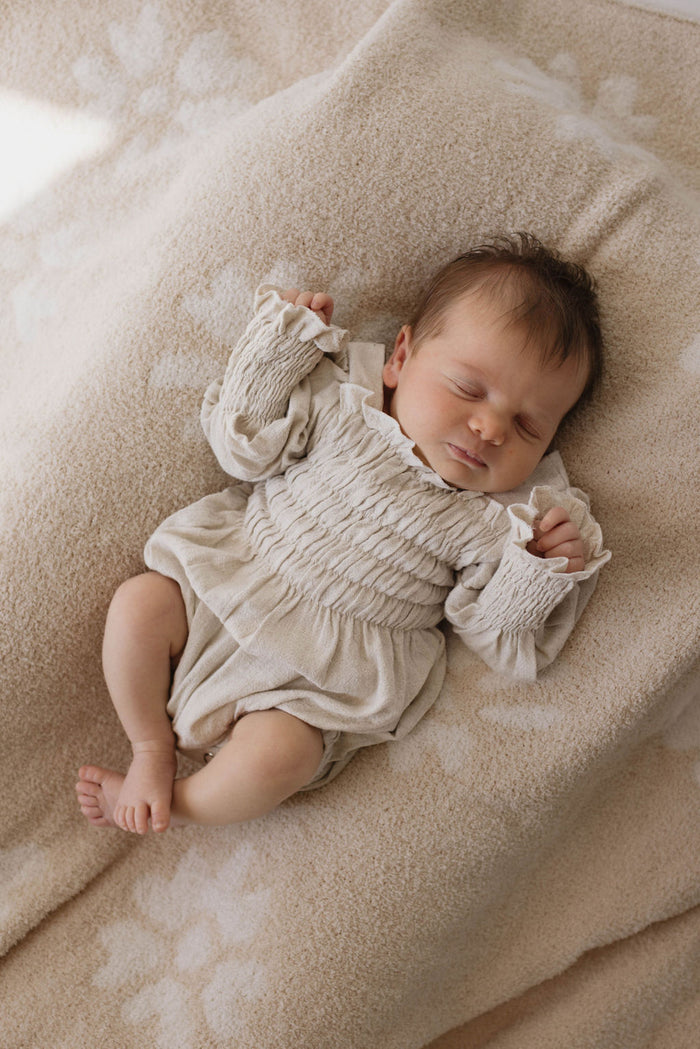 A sleeping baby lies on a Plush Blanket | Lazy Daisy from forever french baby. The soft, beige blanket has a beautiful floral pattern. Dressed in a light-colored onesie made with Microfiber feather yarn and adorned with long sleeves and ruffled details at the collar and wrists, the baby with short, dark hair is peacefully resting.