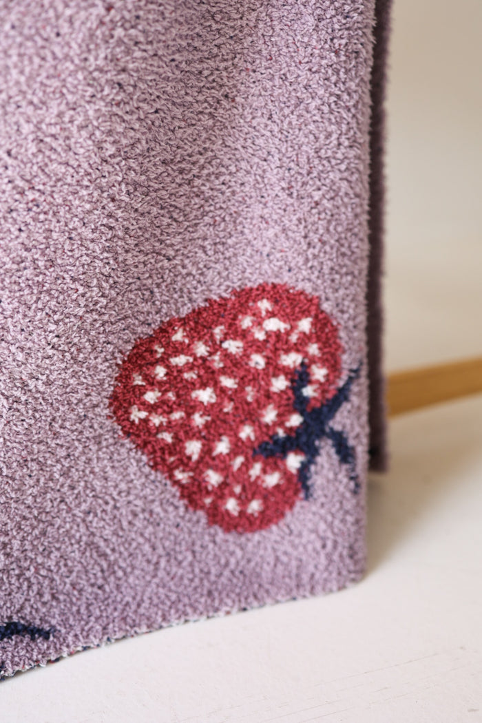 A close-up of the forever french baby Plush Blanket | Matilda in light purple velvet, featuring red strawberries with white seeds and dark stems. The fabric is draped, with a wooden rod partly visible in the background.