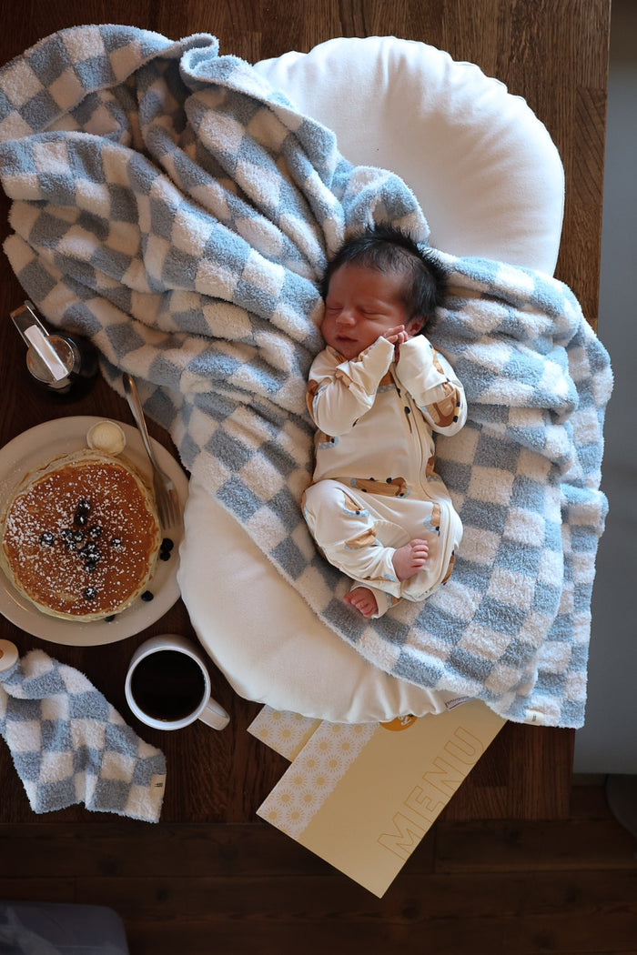 A newborn sleeps on the forever french baby Plush Blanket | Morning Check, featuring a blue-and-white check pattern, next to pancakes, syrup, butter, coffee, and a partially visible yellow menu on a wooden table.