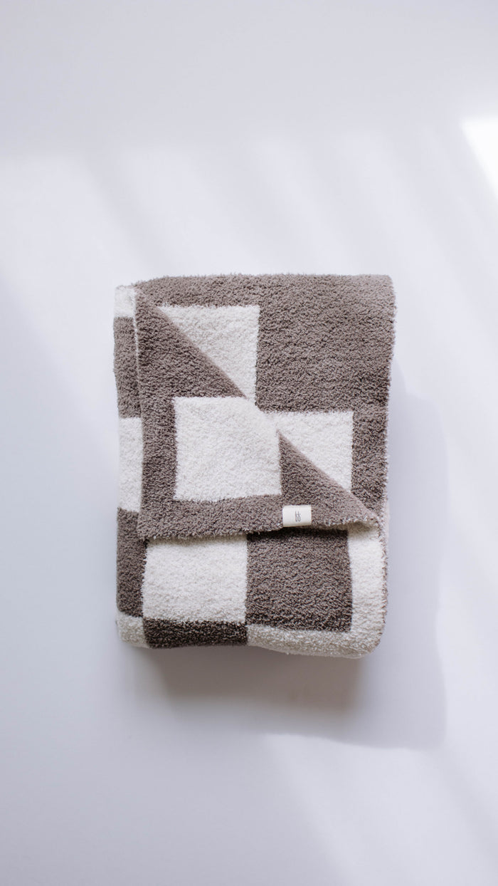A folded, plush blanket labeled as the "Mushroom Checkerboard" by forever french baby lies on a flat white surface, its velvety-rich texture enhanced by soft light and shadows, showcasing a geometric pattern of large gray and white squares and triangles.