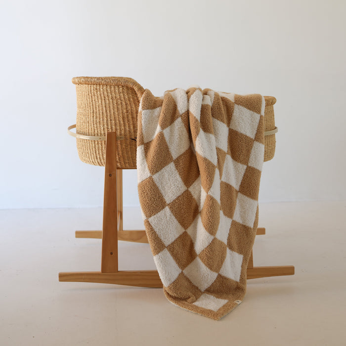 A wicker bassinet on a wooden stand with the forever french baby Plush Blanket | Original Checkerboard (Oeko-Tex certified, beige and white) draped over the side, set against a plain light-colored wall and floor.