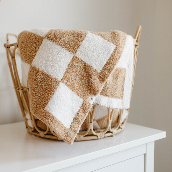 A Plush Blanket | Original Checkerboard by forever french baby, made of OEKO Standard 100 microfiber feather yarn, is draped over the edge of a wicker basket on top of a white dresser. The beige and white checkered pattern, combined with its cozy texture, creates a warm, minimalist feel in the room.