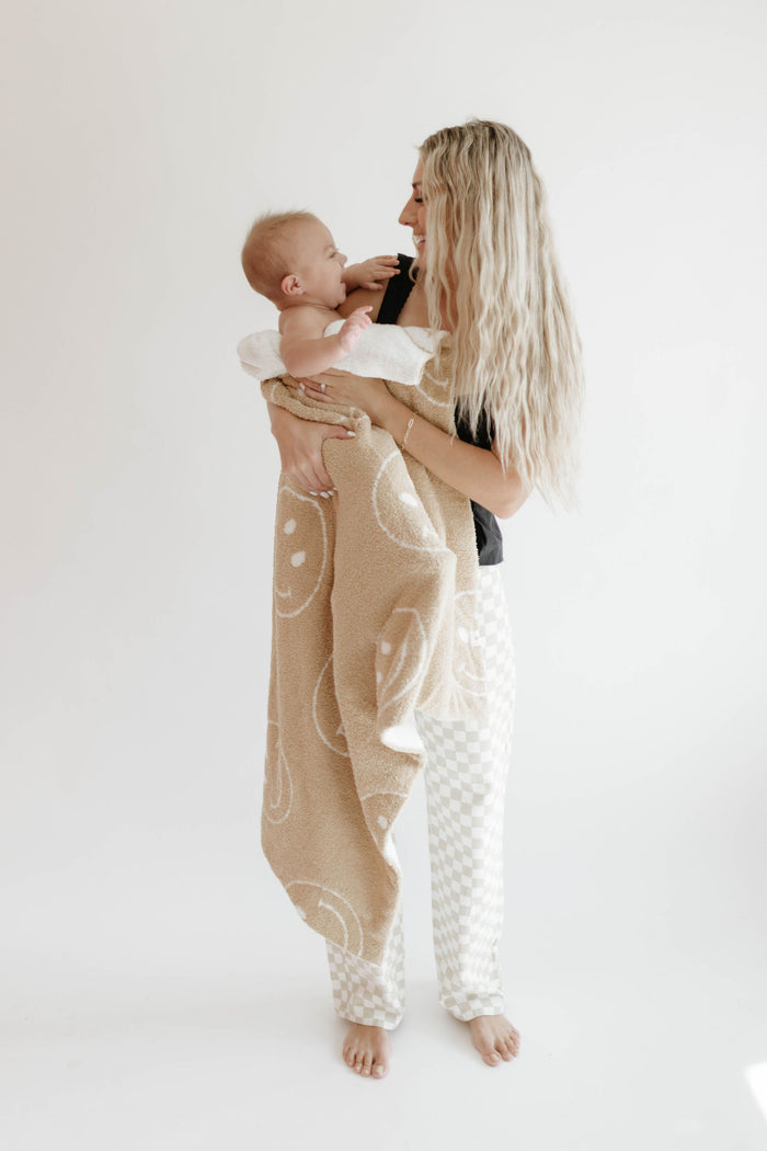 A woman with long blonde hair holds a smiling baby wrapped in the forever french baby Plush Blanket | Original Just Smile, featuring white smiley faces. Barefoot, they stand against a plain white background, looking at each other happily.