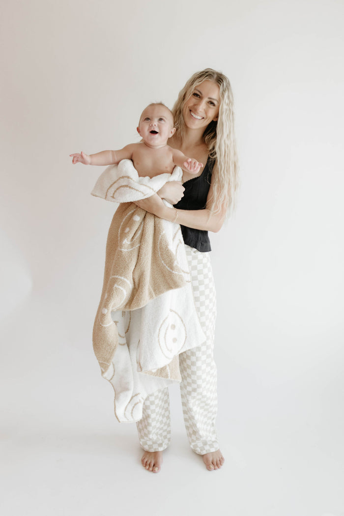 A smiling woman with long blonde hair holds a happy baby wrapped in a forever french baby Plush Blanket | Original Just Smile, featuring smiley faces and soft microfiber feather yarn, against a plain white background.