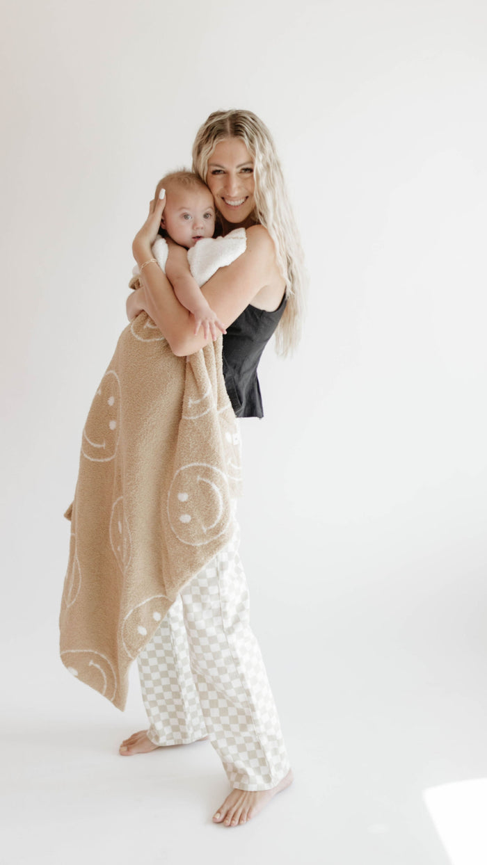 A smiling woman with long blonde hair holds a Forever French Baby brand Plush Blanket | Original Just Smile, a tan blanket with white smiley faces, while barefoot in black top and checkered pants against a plain white background.