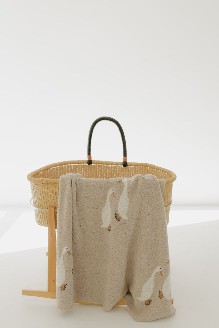 A woven bassinet with black handles sits on a wooden stand. Draped over the edge is the forever french baby Plush Blanket | Silly Goose, crafted from Oeko-Tex certified microfiber and adorned with charming white goose prints.