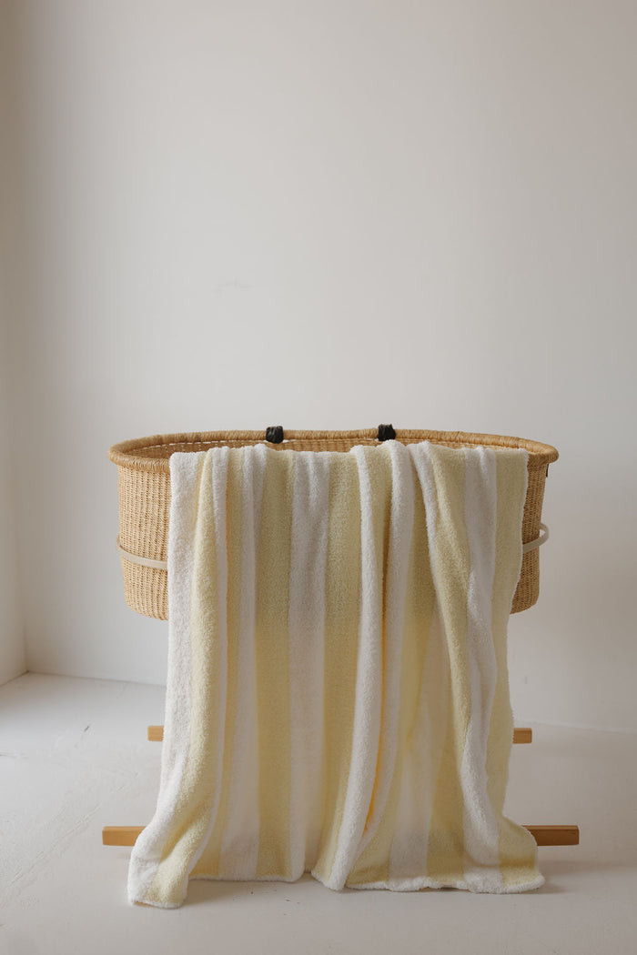 A wicker bassinet on wooden legs with the Plush Blanket | Sunny Stripe by forever french baby—an Oeko-Tex certified, soft baby blanket in striped yellow and white—draped over the side, set against a plain white wall and floor.