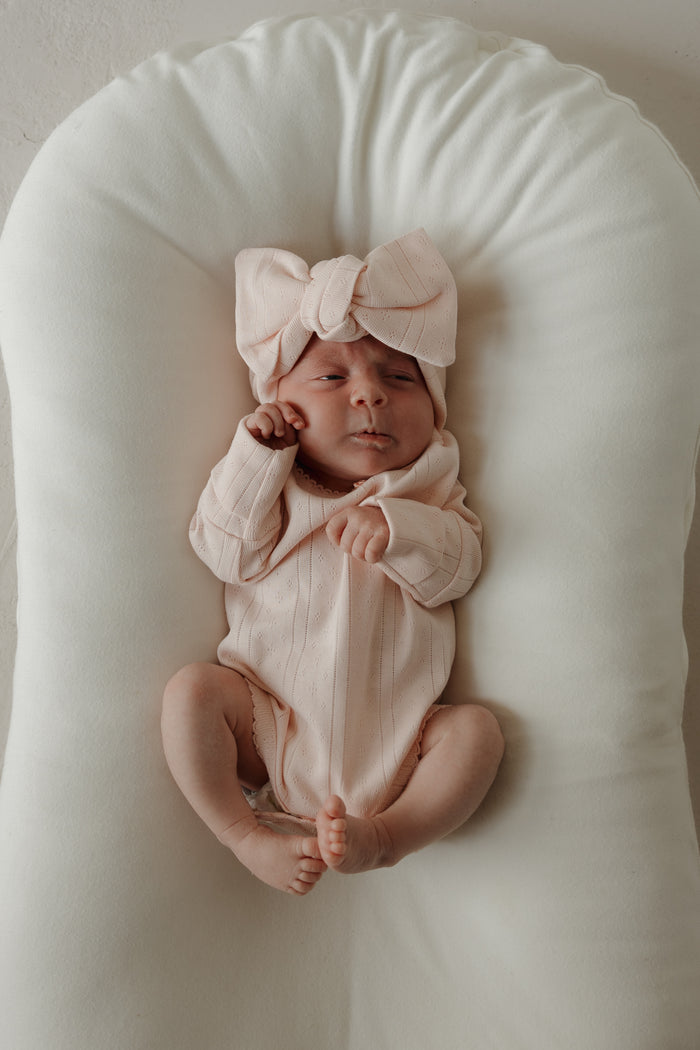 A baby wearing the forever french baby Pointelle Head Wrap in Ballerina and a light pink pointelle cotton outfit lies on a soft, white cushion, gazing upward with a relaxed expression.