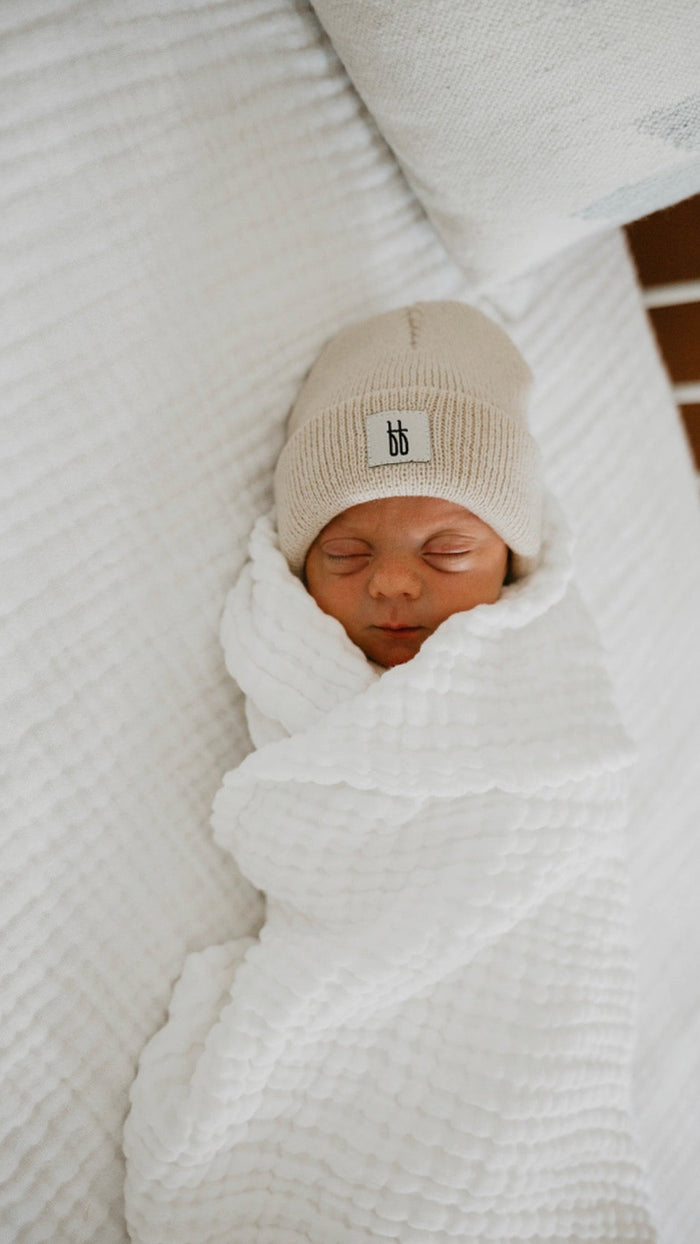 A newborn baby is peacefully sleeping, wrapped snugly in a Forever French Baby Quilt | Cloud. The textured white quilt and the beige knit hat, adorned with a small "bb" tag, perfectly complement each other. Set against a soft white surface, the serene scene suggests washing with like colors.