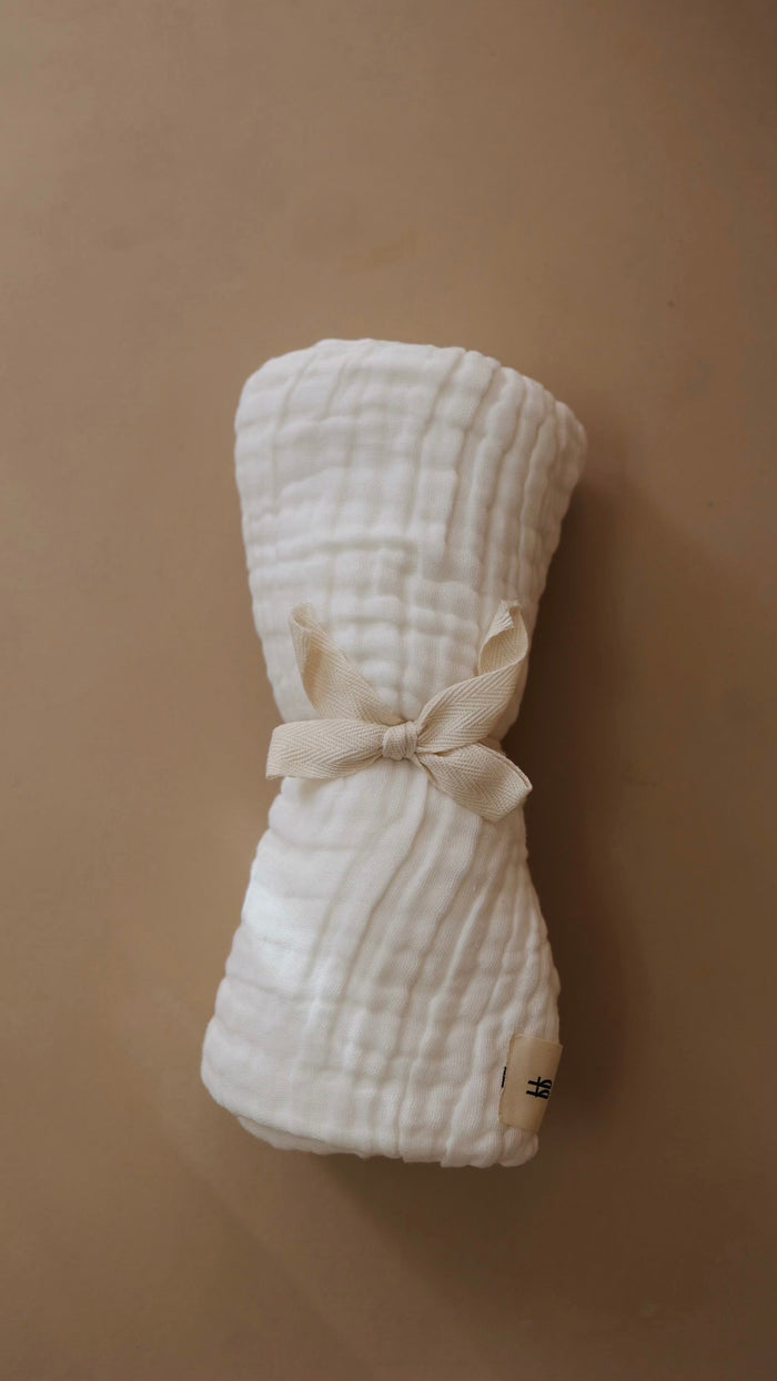 A neatly rolled 40 X 40 white muslin cloth from the Quilt | Cloud collection by forever french baby, tied with a beige ribbon, is placed on a brown surface. The fabric features a textured, quilted appearance, and a small rectangular tag marked "forever french baby" is visible at the bottom corner of the roll.
