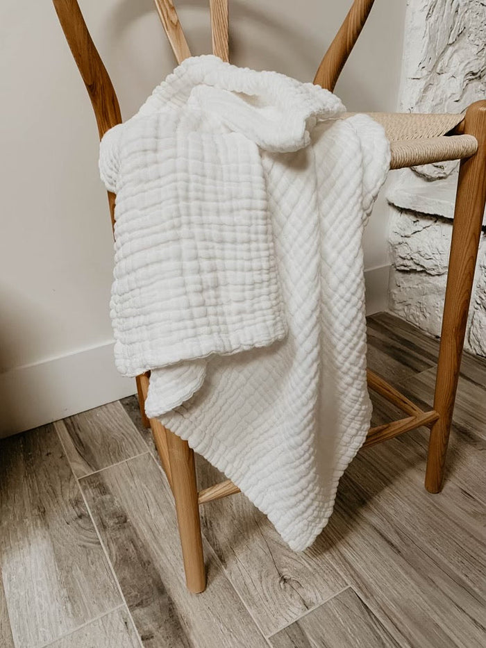 A soft, white, textured Quilt | Cloud from forever french baby is draped over the back of a wooden chair with a woven seat. The chair is positioned near a light-colored wall and stone decor. The flooring features wide, light-colored wood planks. For best care, wash with like colors.