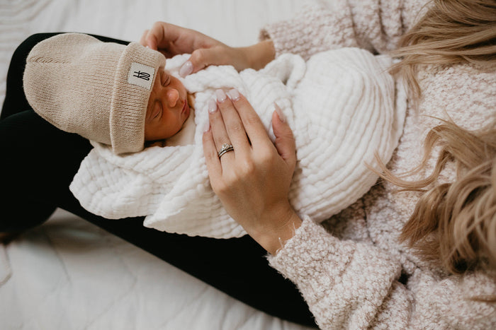 A newborn baby dressed in a beige knit hat is wrapped in a forever French baby's Quilt | Cloud and cradled in a woman's arms. The woman, wearing a fluffy sweater, gently holds the baby, showcasing her manicured nails and a ring on her finger.