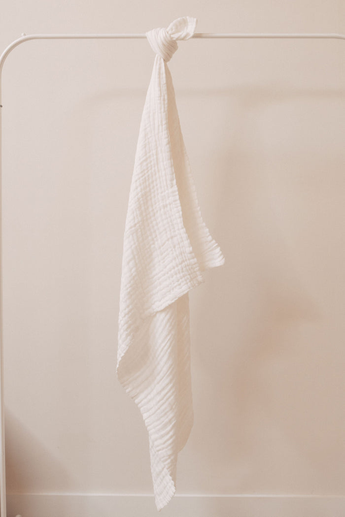 A Quilt | Cloud from the forever french baby collection, featuring a white, textured, gauzy fabric, is hanging on a minimalist metal rack against a light-colored wall. Measuring 40 X 40 inches, the quilt is draped unevenly, with one end hanging longer than the other for a casual and relaxed appearance. Remember to wash with like colors.