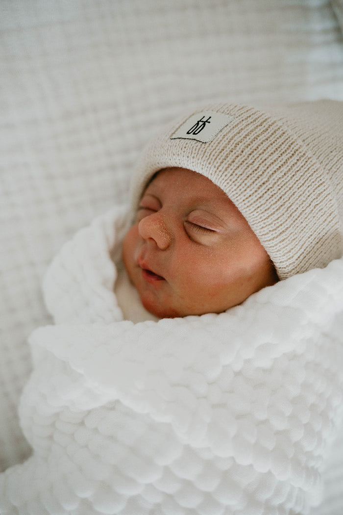 A newborn baby sleeps peacefully, wrapped in a soft white Quilt | Cloud from the forever french baby collection. The baby wears a beige knit hat with a small embroidered design. The background is a textured off-white fabric. Quilt size 40 x 40 inches, wash with like colors for best care.