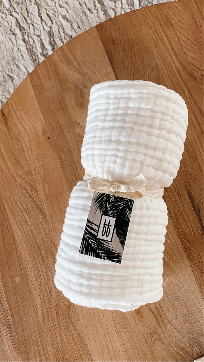 A rolled-up, white, textured Quilt | Cloud is placed on a wooden surface. The quilt is tied with a beige ribbon and features a tag showcasing black palm leaves and the letters "bb" in white. Measuring 40 X 40, this Forever French Baby quilt should be washed with like colors. A portion of a beige, textured rug is visible in the background.
