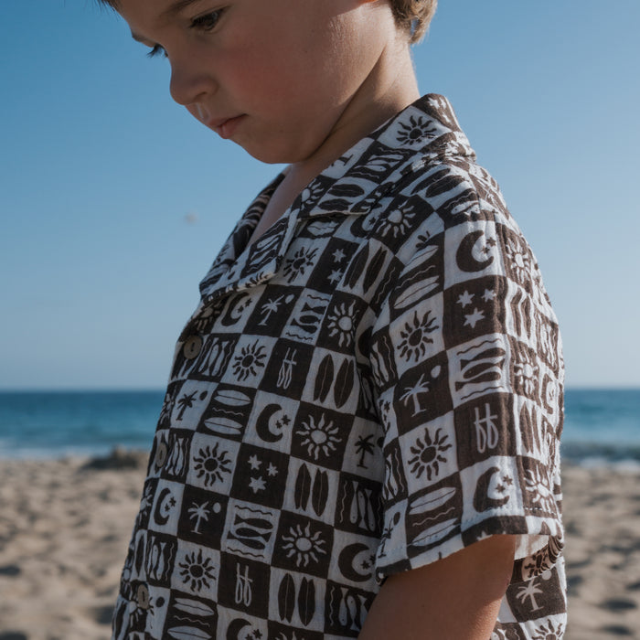 A young child wears the forever french baby Short Sleeve Muslin Button Up in Summer Check on a sandy beach, gazing down with the blue sky and ocean as a backdrop.