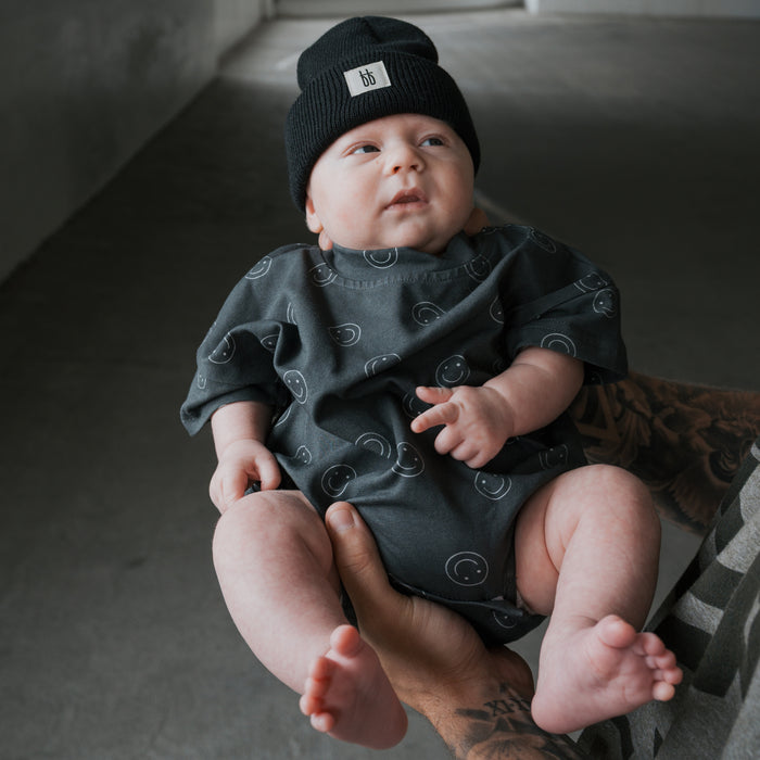 A baby wears the forever french baby Short Sleeve Romper | Smiley and a black beanie, held by a tattooed adult in a gray room.