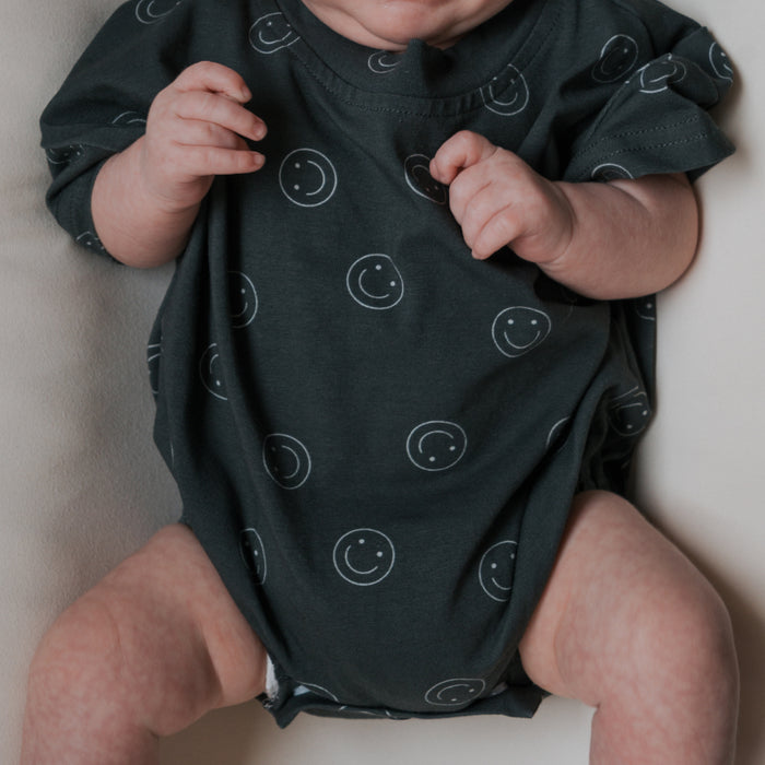 A baby in the forever french baby Short Sleeve Romper | Smiley, featuring a dark cotton fabric with white smiley face patterns, lies on a light surface with arms slightly raised and relaxed legs. The baby's face is partially out of frame.
