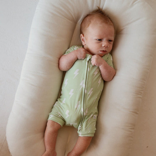 A light-skinned, red-haired baby lies on a soft cream cushion in the forever french baby Shortie Bamboo Zip | Flash romper—a light green bamboo outfit with white lightning bolts—looking to the side with hands near the zipper.