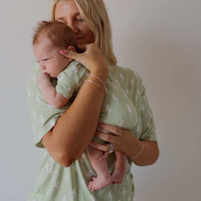A blonde woman in a green lightning bolt-patterned outfit holds a newborn dressed in forever french baby’s Shortie Bamboo Zip | Flash pajamas, perfectly matching her. The background is plain and light-colored.