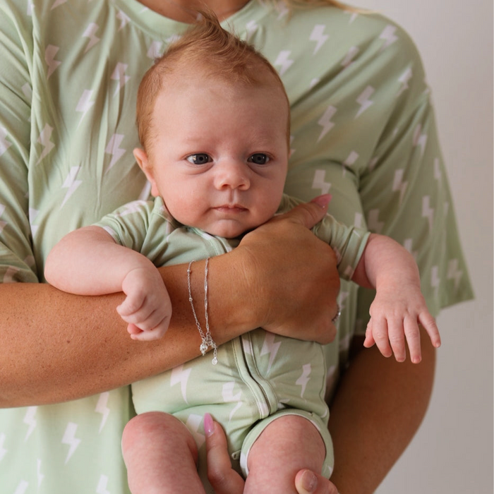 A baby with reddish hair is calmly held by an adult, both wearing forever french baby Shortie Bamboo Zip | Flash pajamas in light green with white lightning bolts, as the adult gently supports the baby.
