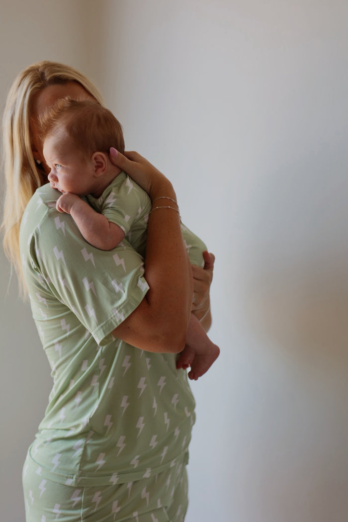 A woman wearing forever french baby Shortie Bamboo Zip | Flash pajamas in light green with white lightning bolts holds a baby in matching hypoallergenic clothing, standing against a plain, light background as the baby rests on her shoulder.