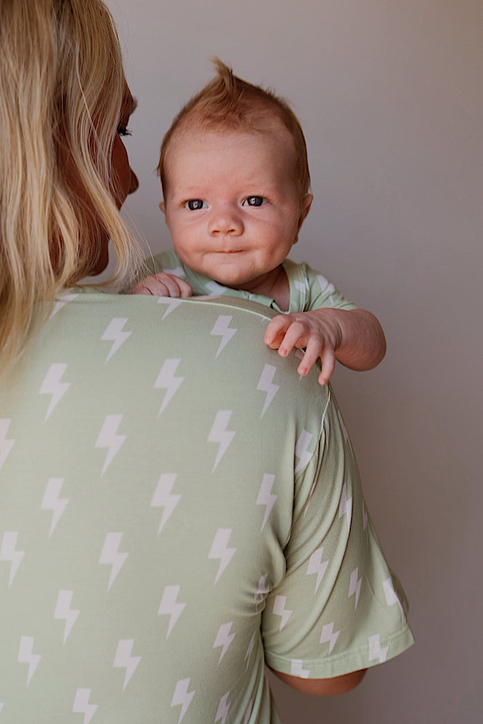 A baby with reddish-blond hair smiles while resting on an adult’s shoulder. Both wear matching forever french baby Shortie Bamboo Zip | Flash pajamas—light green, bamboo fabric, adorned with white lightning bolt patterns.