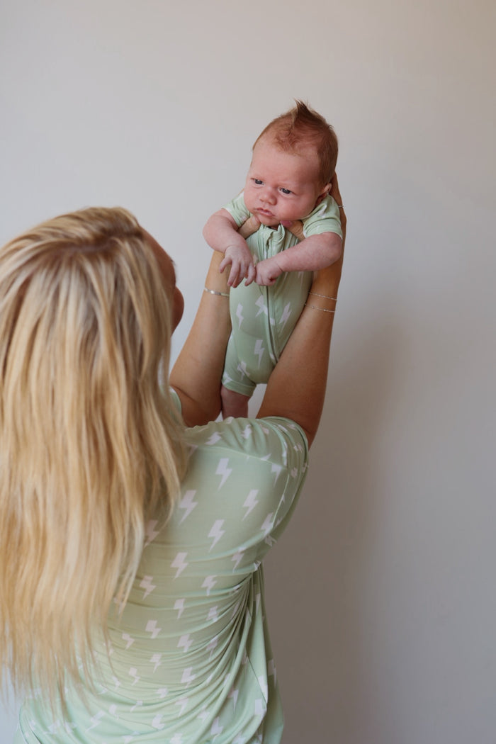 A blonde woman holds a baby facing her, both wearing forever french baby's Shortie Bamboo Zip | Flash pajamas in light green with white lightning bolts. The plain background highlights their soft, hypoallergenic clothing.