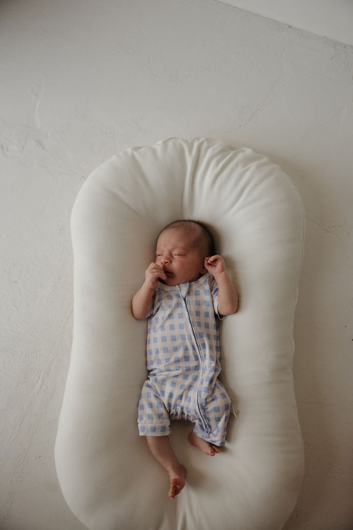 A newborn baby in forever french baby’s Shortie Bamboo Zip | Marilyn—blue and white checkered, with eyes closed and hand near mouth—rests on a white cushioned lounger against a light background, perfectly cozy in hypoallergenic clothing designed for delicate skin.