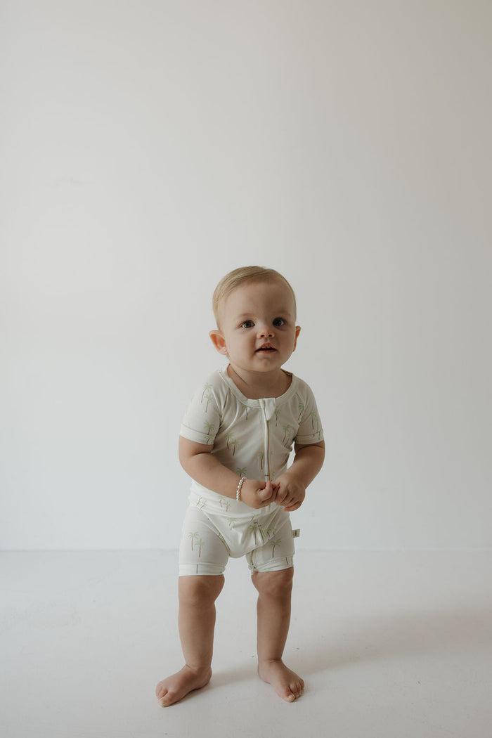 A toddler with short blonde hair stands barefoot on a white floor, wearing the forever french baby Shortie Bamboo Zip in the Palm print—a light-colored, hypoallergenic romper. The background is plain white, and the child gazes slightly upward with a neutral expression.