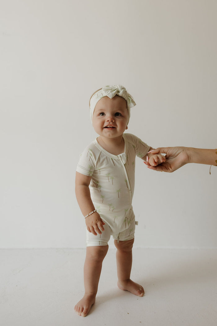 A baby wearing the forever french baby Shortie Bamboo Zip in the Palm print stands barefoot and smiling, holding an adult’s hand against a plain white background.